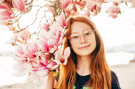 Spring portrait of adorable red-haired preteen kid girl with magnolia flowers, child wearing eyeglasses