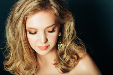 Close up portrait of beautiful young woman with blond hair and professional make up, posing on black background, looking down
