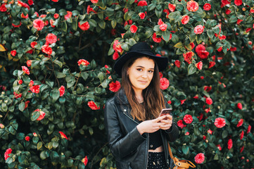 Outdoor portrait of beautiful young woman wearing black leather jacket and hat, posing in camelia...