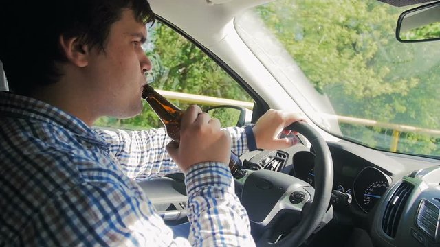 Slow motion footage of drunk man drinking beer from bottle while driving a car. It is dangerous and may cause road accident