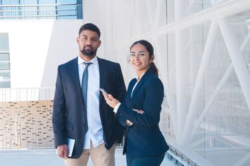 Colleagues posing, holding tablet and smartphone outdoors. Business man and woman looking at camera with building wall and railing in background. Business people and break concept. Front view.