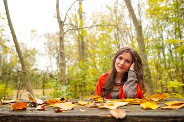 Beautiful romantic girl in park autumn scenery, sitting down at a wooden table covered with yellow leaves, looking at the camera. Gorgeous young woman outdoors. Close-up landscape shot, natural light