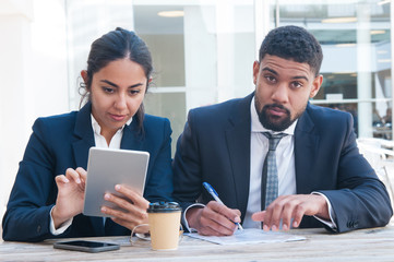 Serious colleagues using tablet and working at wooden desk. Business man and woman wearing formal clothes and sitting in office. Business work concept. Front view.
