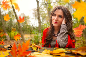 Beautiful romantic girl in park autumn scenery, sitting down at a wooden table and looking away, blurred yellow leaves are falling in the foreground. Close-up shot in natural light, vibrant colors