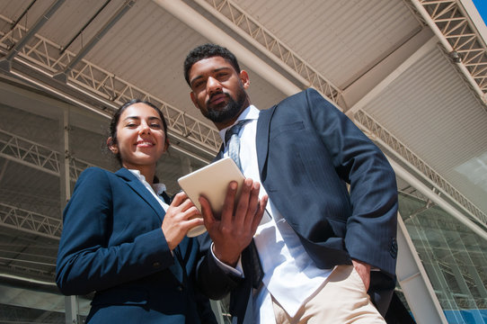 Positive colleagues posing, holding tablet and drink outdoors. Business man and woman looking at camera with building constructions in background. Business people concept. Low angle front view.