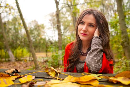 Beautiful Romantic Girl In Park Autumn Scenery, Sitting Down At A Wooden Table Covered With Yellow Leaves, Looking Away. Gorgeous Young Woman Outdoors. Close-up Shot In Natural Light, Vibrant Colors