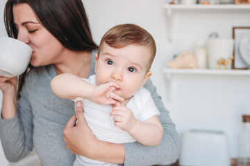 Young mother brunette woman with baby boy in arms drinking tea in white modern kitchen at home