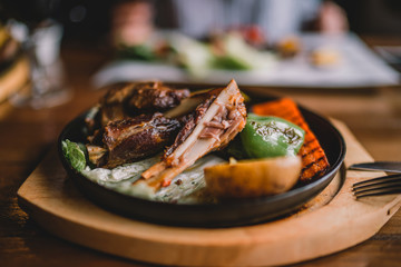 Spicy grilled lamb leg with vegetables and herbs on a roasting cast iron. Barbecue lamb with vegetables. Healthy food. Eating and leisure concept. Woman having dinner at table with food. Toned image.