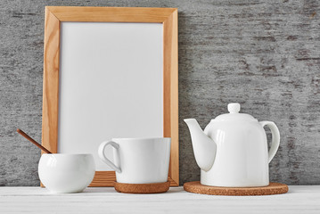 Cup of tea, teapot, sugar bowl and empty frame with copy space on gray background, mockup