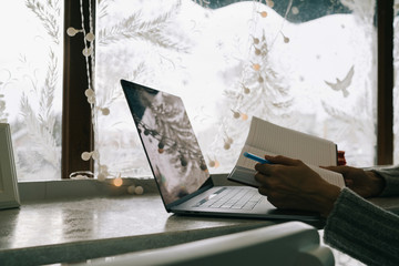 Magical Christmas atmosphere. Paper notebook, laptop and woman hands in front of the winter window. Cozy winter. Morning planning and inspiration. Winter and people concept.