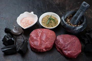 Raw fresh marbled beefsteaks with condiments, charcoal and a decorative grill on a dark brown stone background
