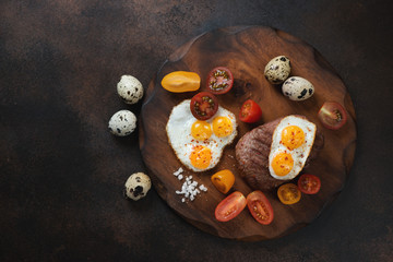Grilled marbled beefsteak in tuscan style - with fried quail eggs and cherry tomatoes. Flatlay on a dark brown metal background with space