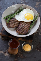 Barbecued beef schnitzels served with fried egg and sauces, vertical shot on a brown stone background
