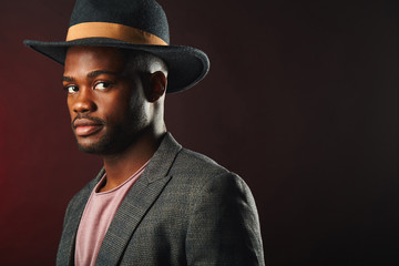 African cnfident business man, dressed in grey formal wear with black hat put on head, posing in studio on dark smoky background