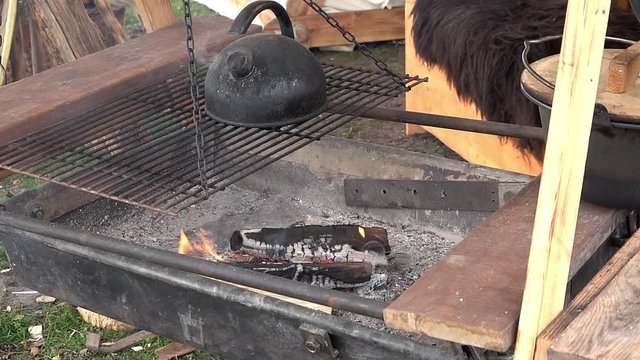 Old water kettle covered with black patina on a grill grate, under which a fire of logs burns