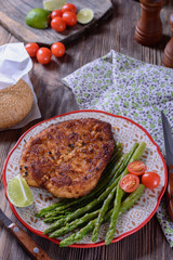 Hot appetizing schnitzel with crispy crust on a plate next to asparagus, lime and cherry tomatoes on a wooden table are ready for eating