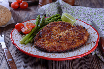 Hot appetizing schnitzel with crispy crust on a plate next to asparagus, lime and cherry tomatoes on a wooden table are ready for eating