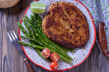 Hot appetizing schnitzel with crispy crust on a plate next to asparagus, lime and cherry tomatoes on a wooden table are ready for eating