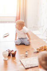 Happy infant boy eats ready-made cakes and having fun with cutting board covered with flour after cake dough sitting on wooden kitchen table. Child s development in home interior