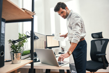 Multitasking. Side view of successful bearded trader or businessman in formal wear working with laptop while standing at his modern office.