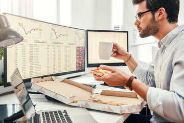 Food for productive work. Side view of young bearded trader in eyewear is eating hot pizza while...