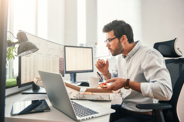 Time for pizza! Side view of young hungry trader is eating hot pizza while looking at monitor screen with financial data in his modern office.