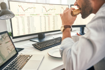 Enjoying lunch. Close-up of bearded trader is eating fresh sandwich and looking at monitor screen with financial data while sitting in his modern office