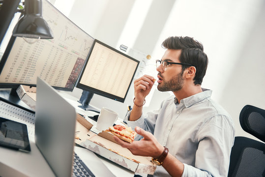 It can not be truth! Disappointed young trader is looking at monitor screen with trading charts and financial data while eating hot pizza in his modern office. - Powered by Adobe