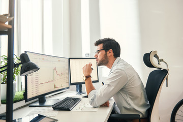 Busy working. Handsome bearded trader in formal wear and eyeglasses is analyzing trading charts and financial data on computer screens while sitting in his modern office.