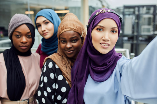 Group of four Asian and African Muslim women in fashionable traditional clothes taking selfie in shopping mall. Focus on white skinned asian beautiful girl in blue dress.