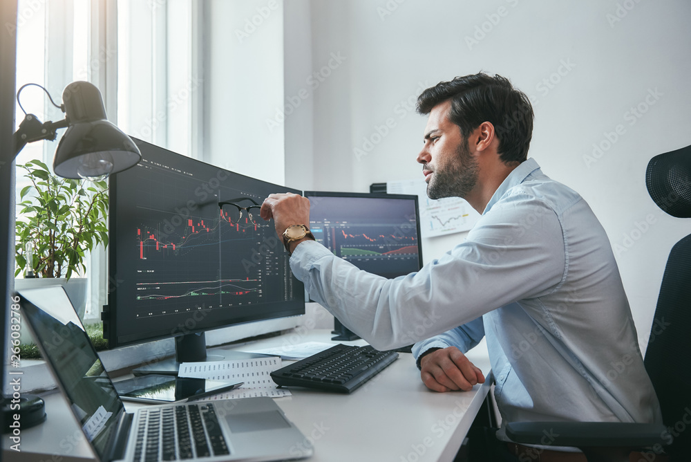Fototapete analyzing data. thoughtful young trader is pointing at graphs on computer screen while working in hi