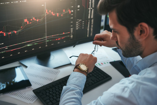 Time to trade. Bearded male trader looking at watch on his hand while working with data and charts on computer screens in his modern office.