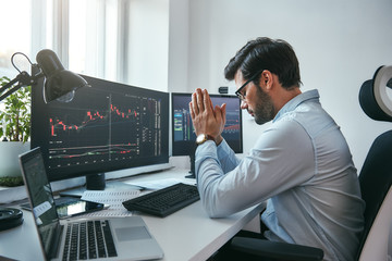 Hoping for the success. Side view of tired young businessman or trader holding palms together while working with data and charts on computers at his modern office.