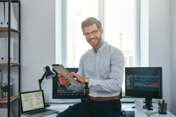 Business success. Cheerful young trader in formal wear is holding digital tablet and looking at camera with smile while standing in front of computer screens with trading charts in the office.