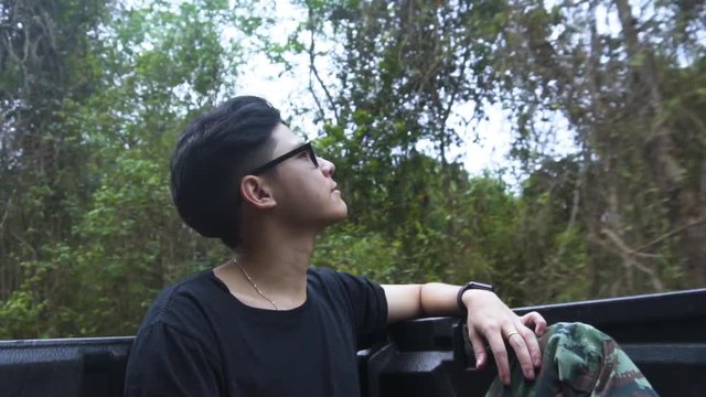 Good looking asian guy sitting at the back of pick-up car and refreshing himself with pure air of forest. slow motion.