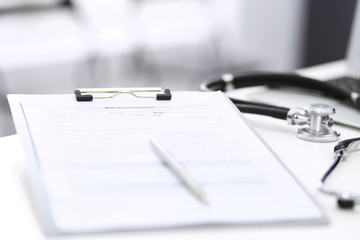 Stethoscope, clipboard with medical form lying on hospital reception desk with laptop computer and busy doctor and patient communicating at the background. Medical tools at doctor working table