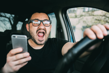 Young man with glasses sitting in his car screaming distracted by smartphone