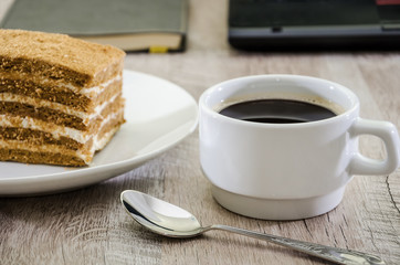 a piece of cake on a plate and a cup of coffee on the background of a laptop and notebook