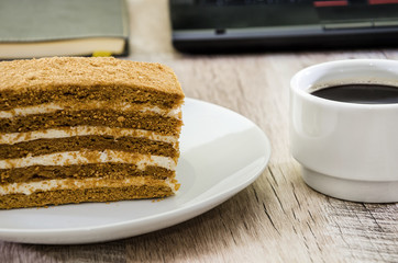 a piece of cake on a plate and a cup of coffee on the background of a laptop and notebook