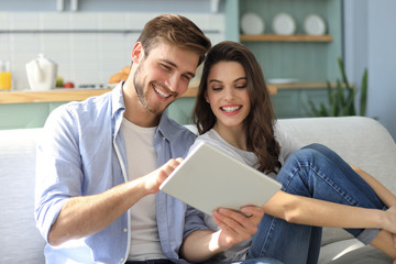 Young couple watching media content online in a tablet sitting on a sofa in the living room. - obrazy, fototapety, plakaty
