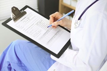 Unknown woman doctor at work at hospital. Pills at medical clipboard. Young female physician write prescription or filling up medical form while sitting in clinic office, close-up of hands