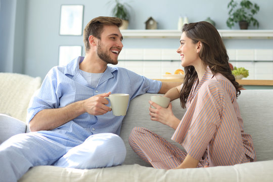 Beautiful young couple in pajamas is looking at each other and smiling on a sofa in the living room.