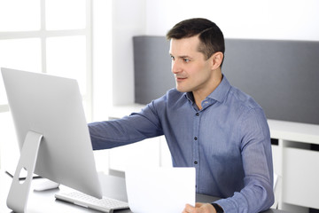 Cheerful smiling businessman working with computer in modern office. Headshot of male entrepreneur or director of a company at the workplace. Business concept 