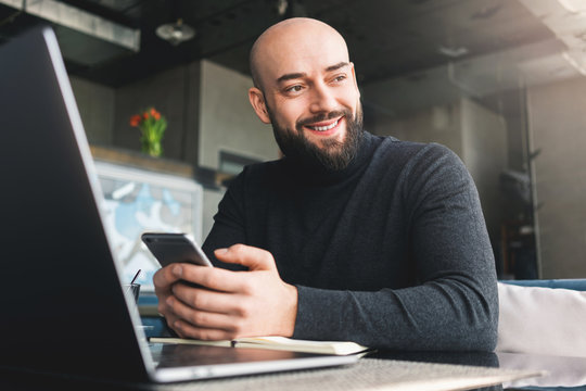 Bald bearded man in black turtleneck working on laptop,holding smartphone while sitting in cafe at table.Freelancer works