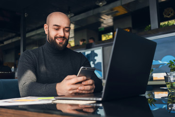 Bald man is working on laptop and uses smartphone while sitting in cafe at table. Freelancer works remotely in coffee shop.
