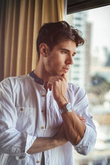 Sexy handsome young man standing and dressing, in his bedroom next to window curtains