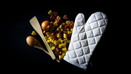 Pasta, cucchiaio di legno, uovo, cipolla e guanto da forno su sfondo nero