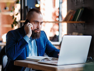 Serious bearded businessman conducts telephone conversations on a mobile phone at the computer. Unhappy man scolds on the phone. Emitations of rage in the office at the manager