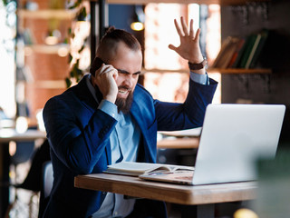 Serious bearded businessman conducts telephone conversations on a mobile phone at the computer. Unhappy man scolds on the phone. Emitations of rage in the office at the manager