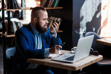 Serious bearded businessman conducts telephone conversations on a mobile phone at the computer. Unhappy man scolds on the phone. Emitations of rage in the office at the manager